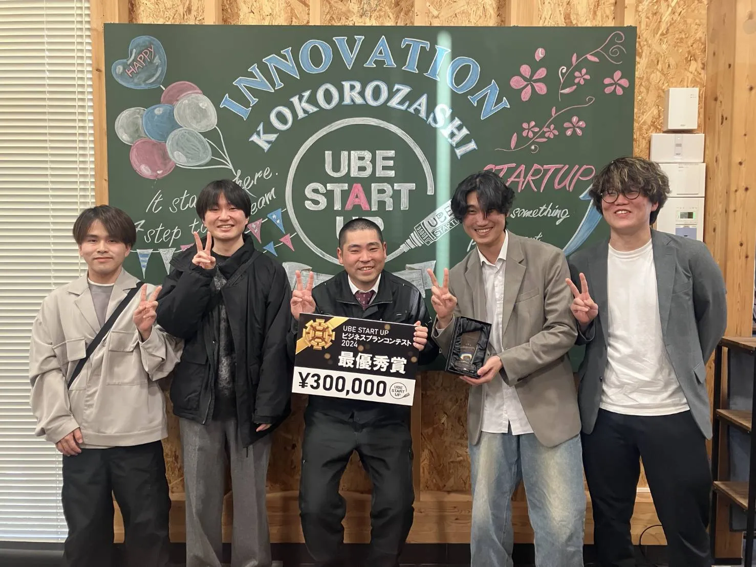 UBE STARTUP ビジネスプランコンテストの最優秀賞の表彰ボードを持って並ぶ5名の職員。