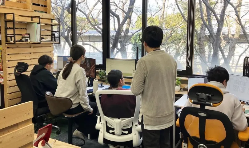 事務所で、窓際に並んだ机に向かって数名の職員が作業している様子。